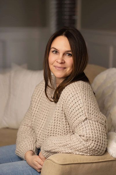 Woman in a beige sweater sitting on a couch, smiling softly in a cozy living room.