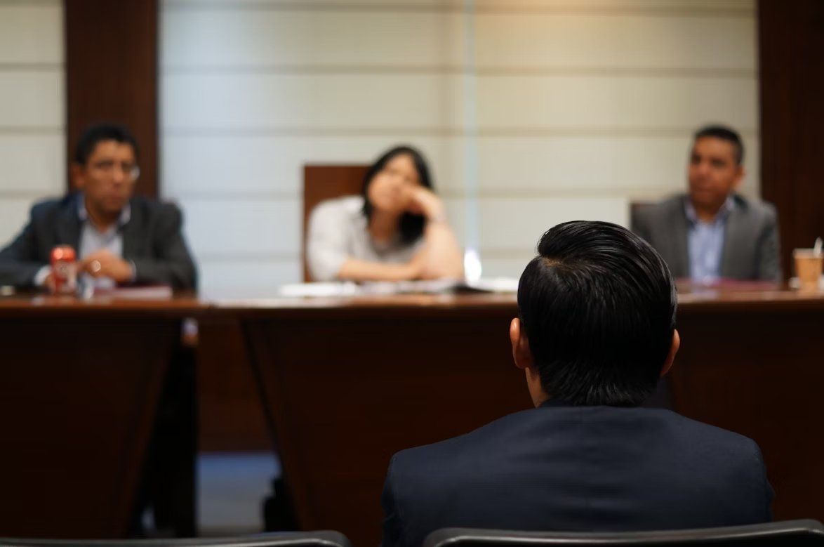 Three people seated at a conference table in a meeting room, seen from behind one attendee.