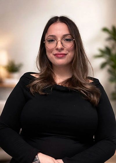 Woman with long brown hair and glasses, wearing a black top, seated and smiling against a softly blurred background
