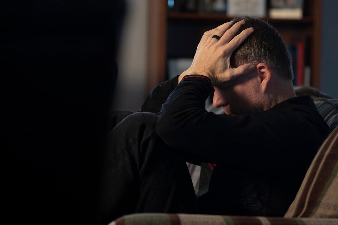 Man seated on a couch, head in hand, appearing stressed in a dim room