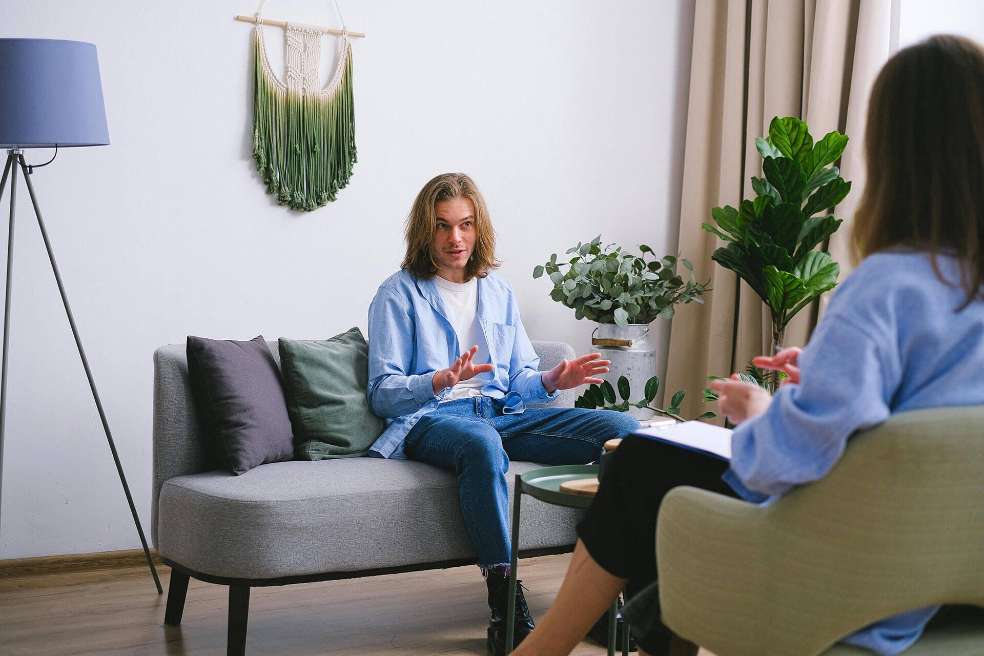 Two people talking in a bright living room, one seated on a gray sofa and one in a chair with a notebook.