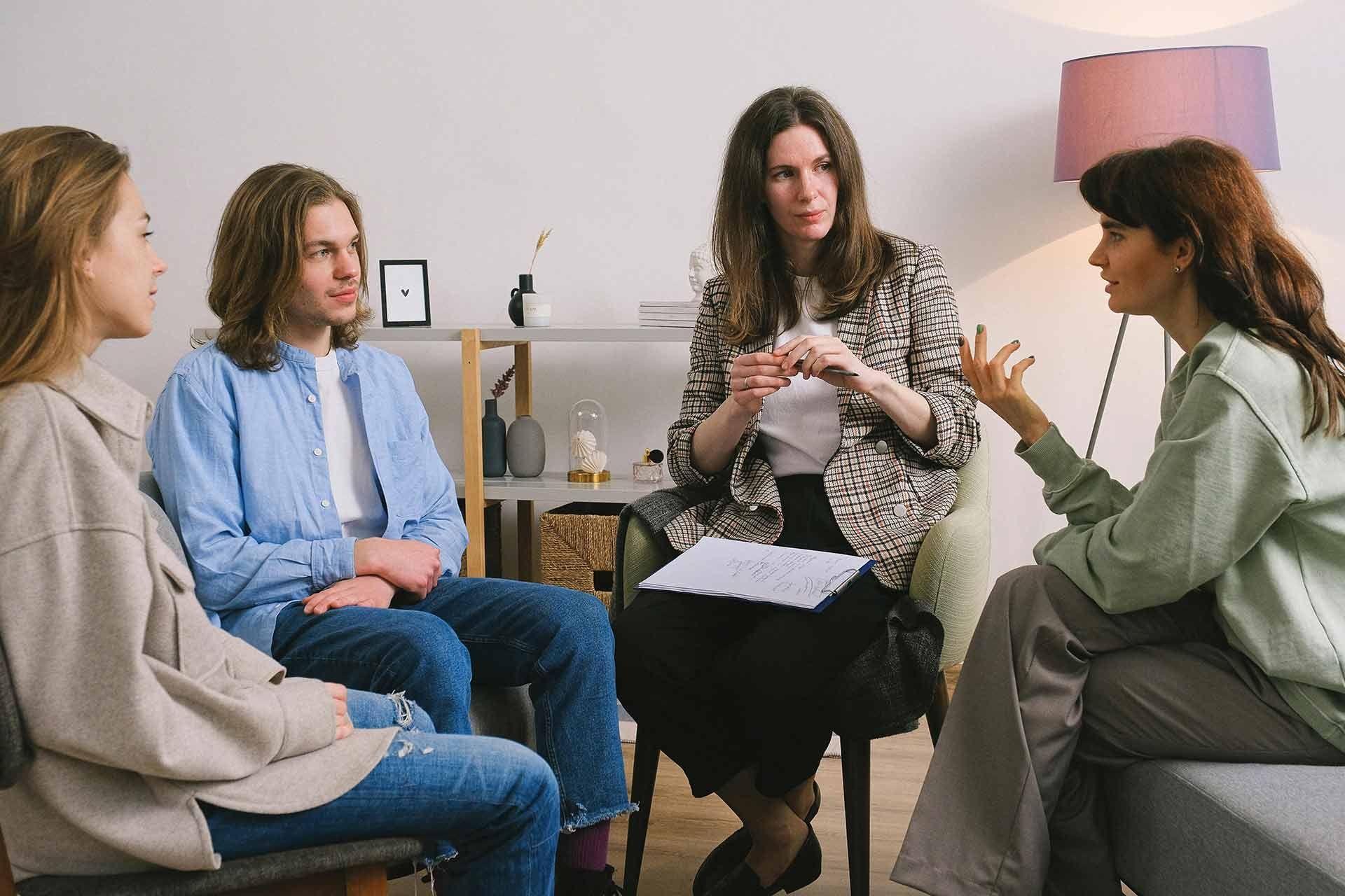 Four people seated in a counseling circle, talking in a bright living room with a floor lamp and side table.