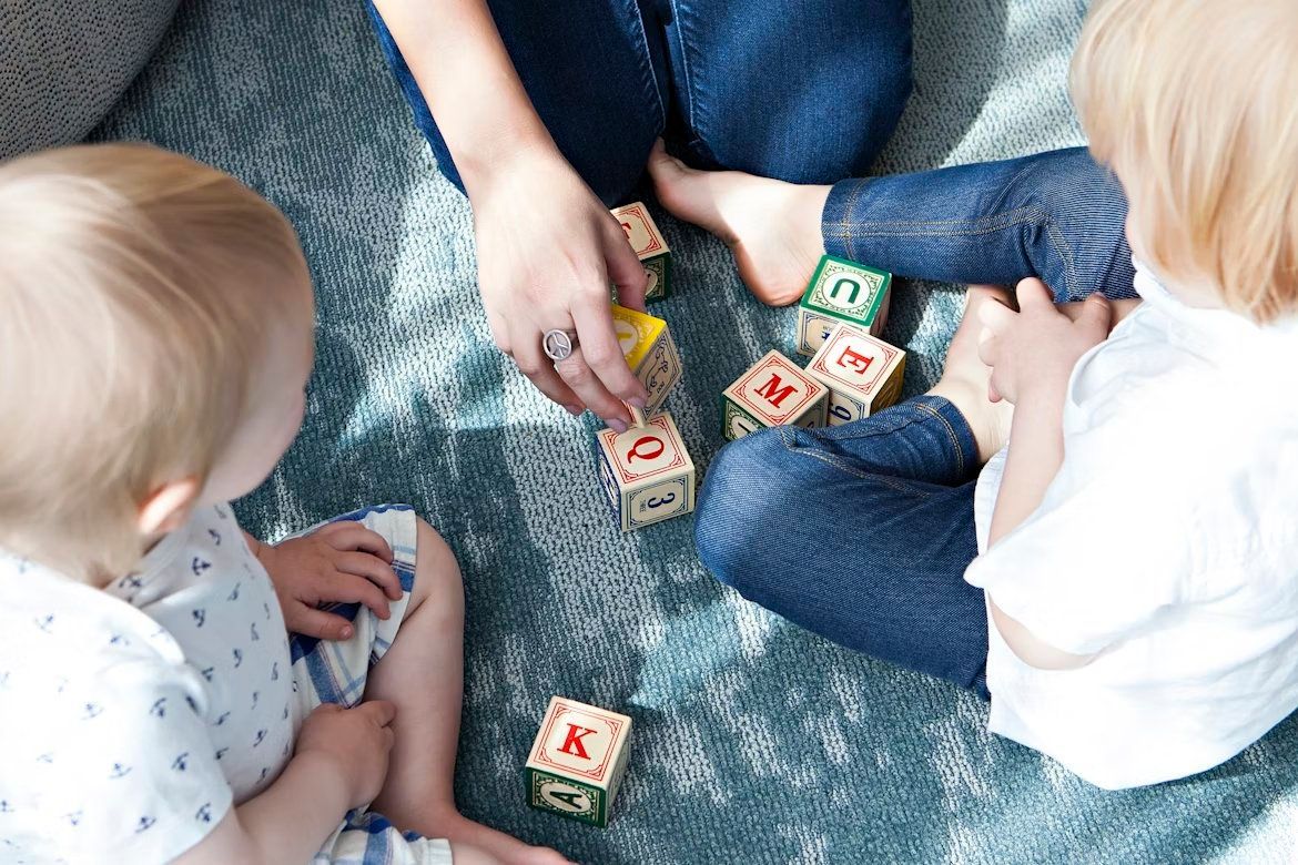 Children playing with wooden letter blocks on a blue rug with an adult nearby
