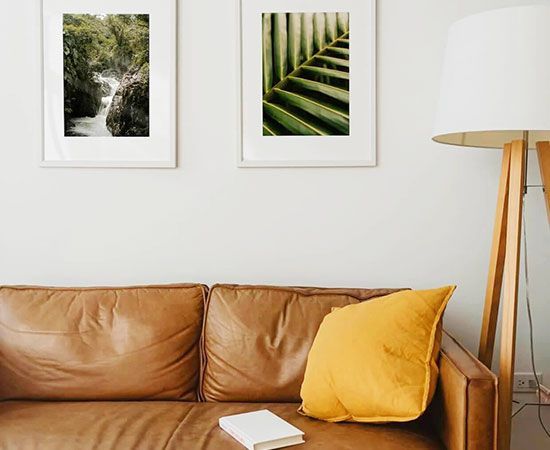 Tan leather sofa with yellow pillow beneath two framed nature prints beside a floor lamp