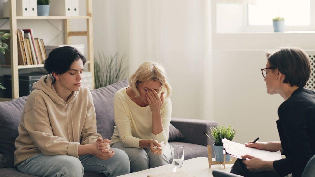 Three people in a bright living room talking with a counselor, one person covering their face in distress.