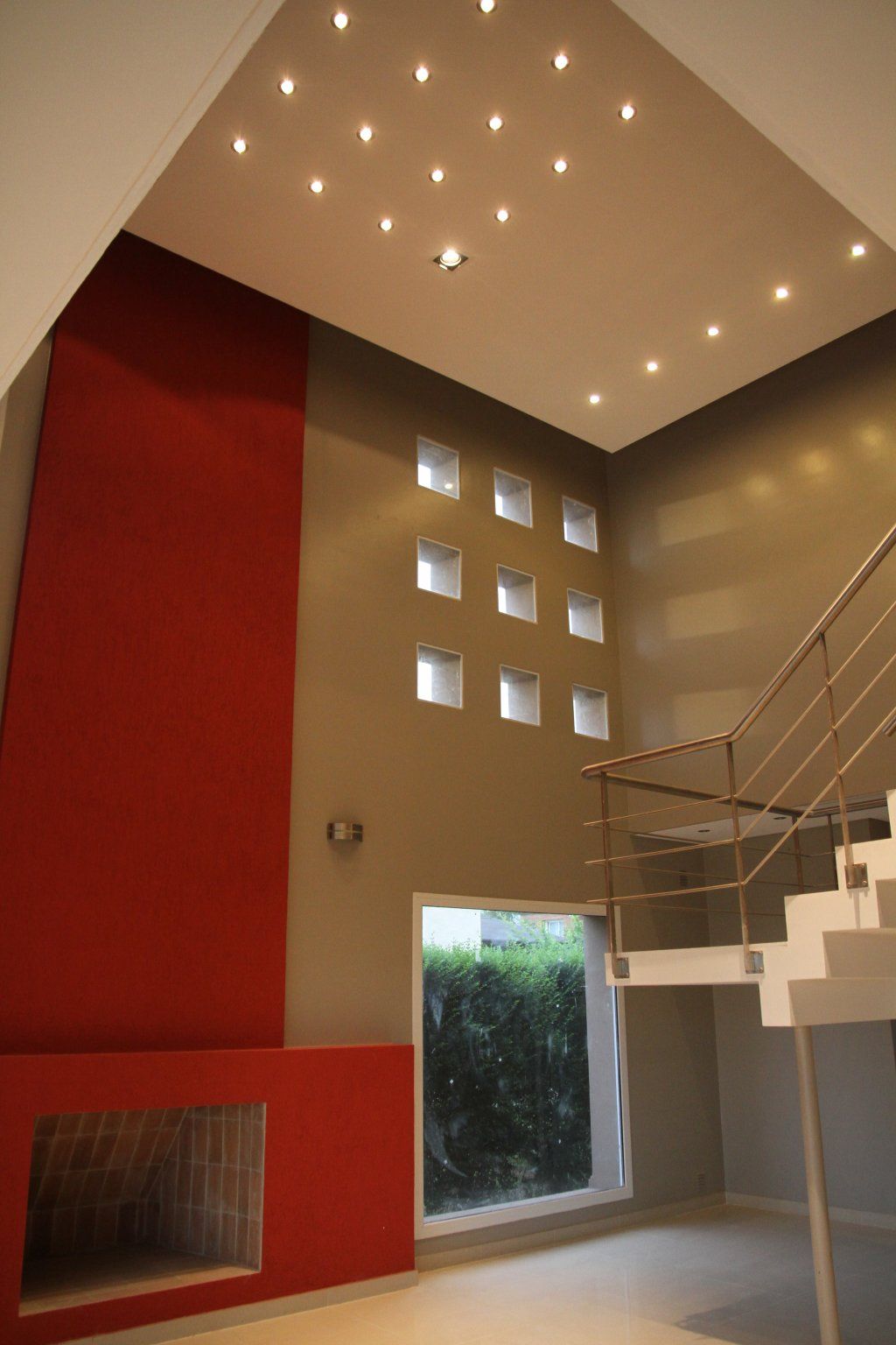 Interior moderno con pared decorativa roja, pequeñas ventanas cuadradas, escalera y luces empotradas en el techo.