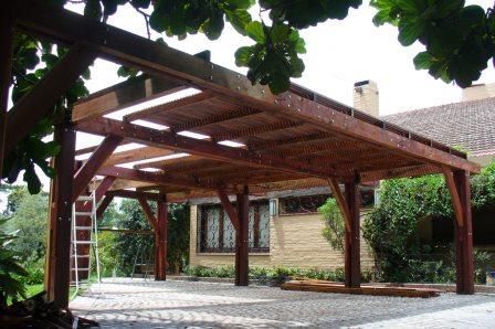 Pérgola/cochera de madera en un patio pavimentado junto a una casa de ladrillo, enmarcada por frondosos árboles.