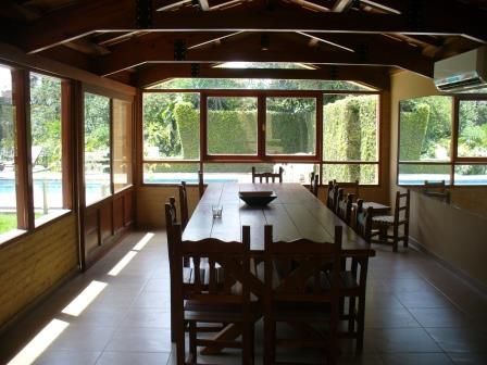 Comedor exterior cubierto con una larga mesa de madera, sillas y grandes ventanales con vistas a la vegetación.