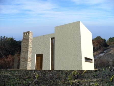 Casa moderna de color beige con una alta chimenea de piedra en un campo de hierba bajo un cielo azul.