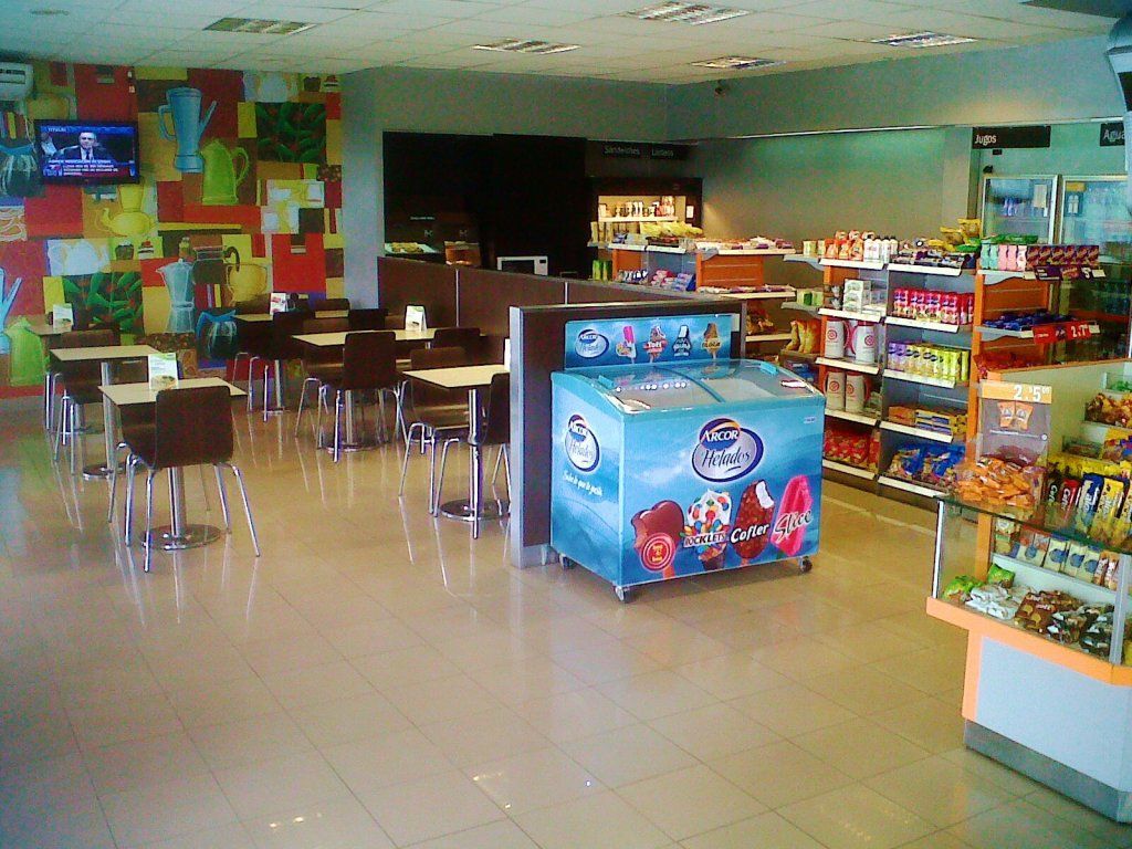 Cafetería vacía con mesas, paredes coloridas y estantes llenos de aperitivos y bebidas.
