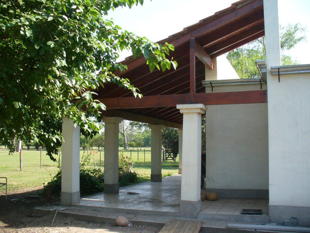 Patio exterior cubierto con columnas blancas, suelo de baldosas y techo rojo inclinado, junto a un jardín con césped.