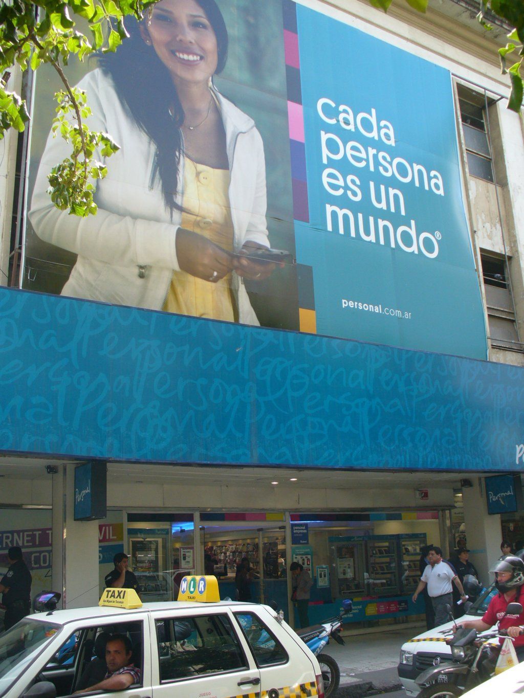 Fachada de tienda con una gran valla publicitaria azul que dice “Cada persona es un mundo” sobre una escena callejera.