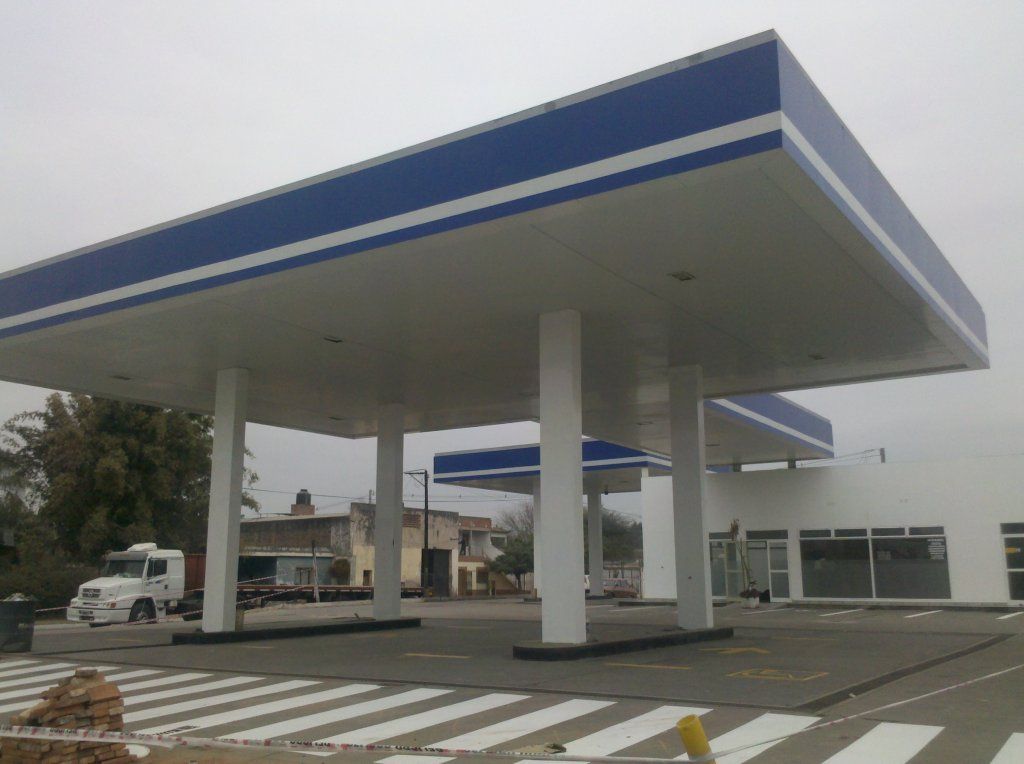 Marquesina vacía de una gasolinera con ribete azul sobre un estacionamiento a rayas bajo un cielo nublado.