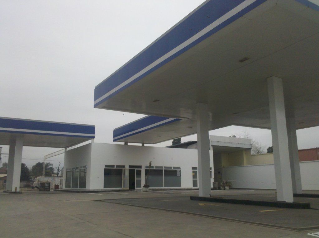 Gasolinera vacía con marquesina ribeteada de azul y edificios blancos bajo un cielo nublado.