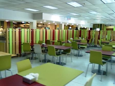 Cafetería vacía con mesas y sillas verdes en una sala luminosa separada por paneles de rayas de colores.