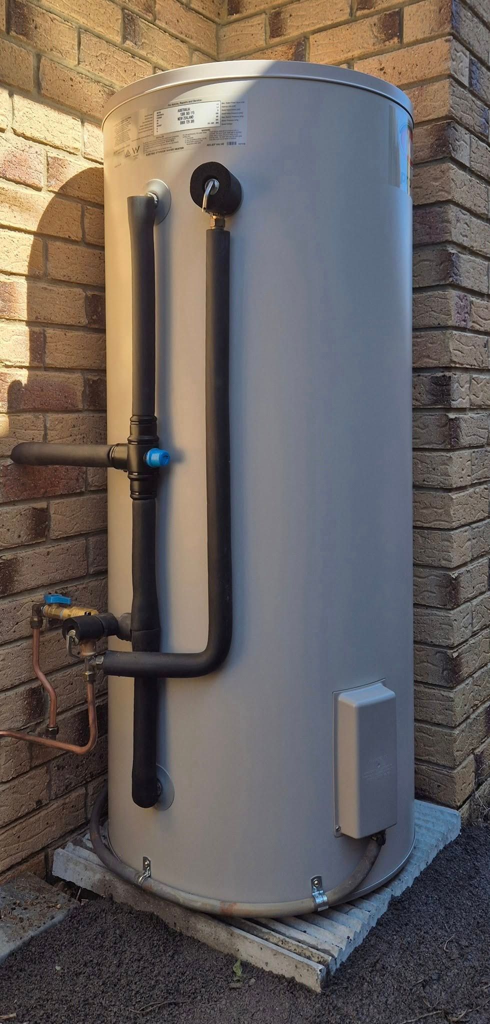 A tall, cylindrical water heater against a brick wall. It has black pipes and sits on a metal base. — 24/7 Hot Water in Gympie, QLD