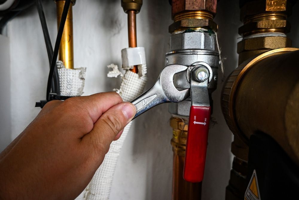 Hand using a wrench to turn a valve on a pipe, indoors. Red handle on valve, copper and brass pipes. — 24/7 Hot Water in Valdora, QLD