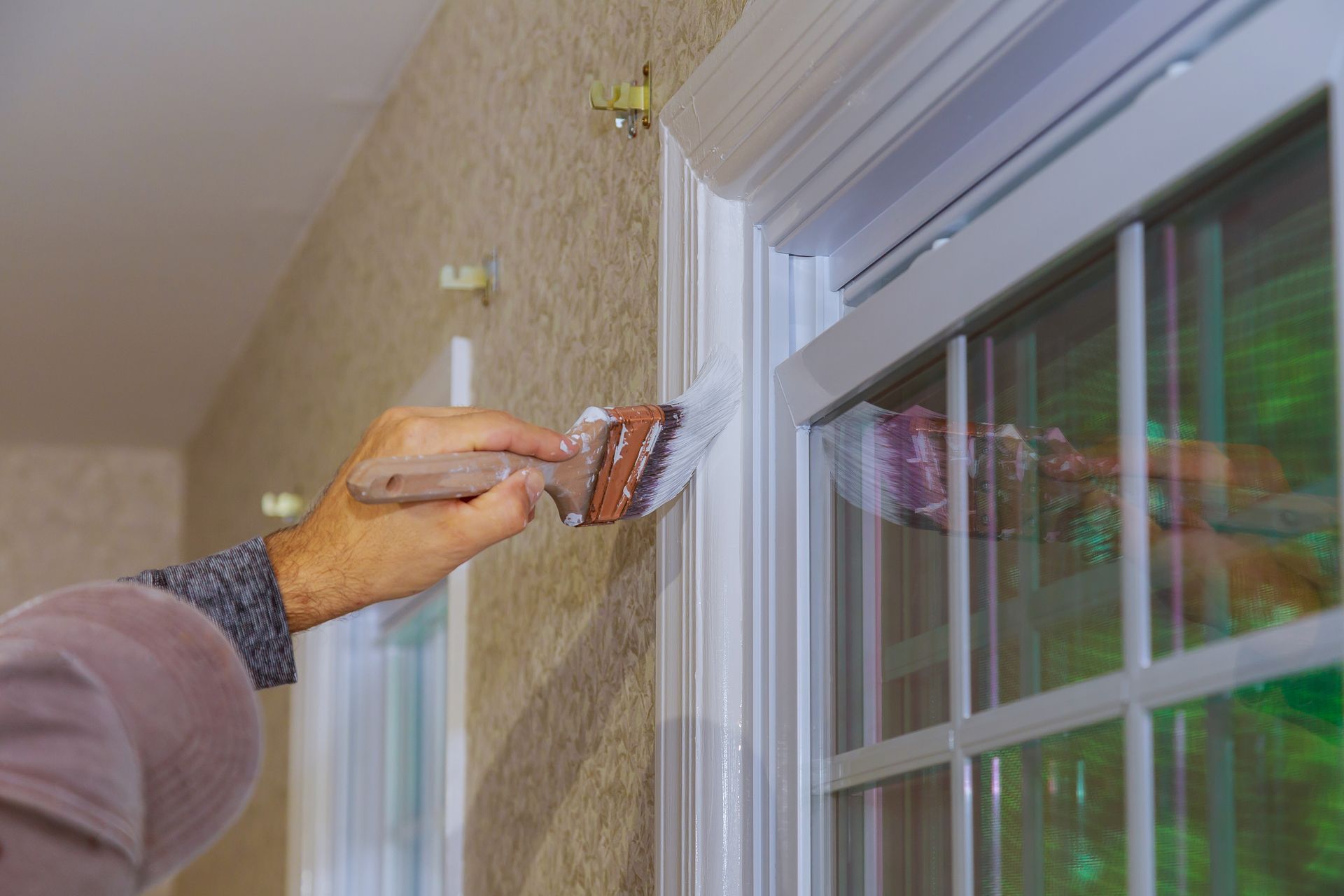 A hand holds a paintbrush, applying white paint to the trim around a window with muntins set into a textured wall. A hand holds a paintbrush, applying white paint to the trim around a window with muntins set into a textured wall.