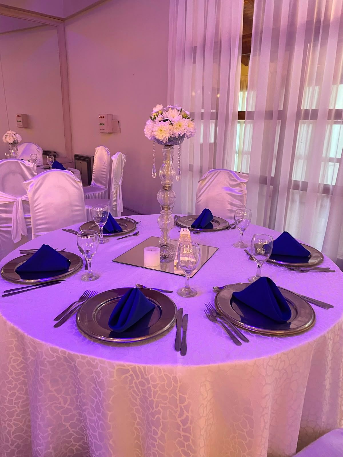 Mesa redonda para banquetes con manteles blancos, servilletas azul oscuro y centro de mesa floral bajo iluminación púrpura.