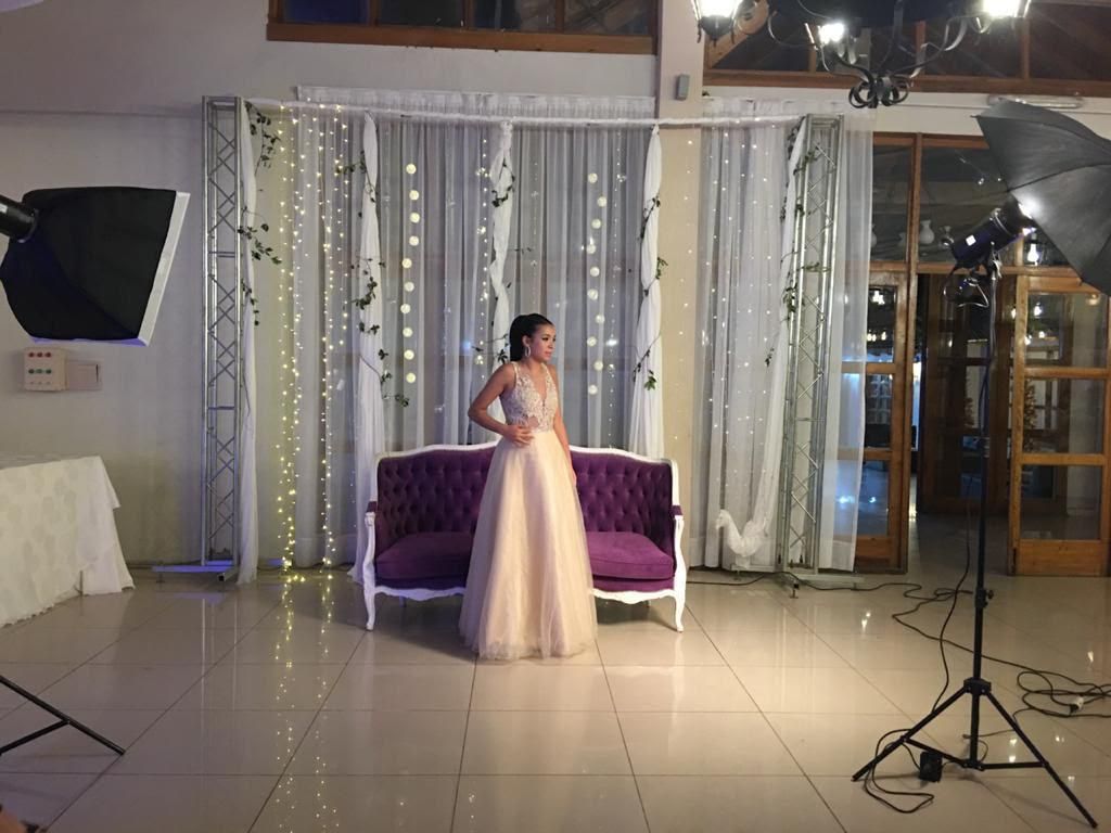 Mujer con un vestido rosa de pie sobre un escenario interior decorado, junto a un sofá morado.