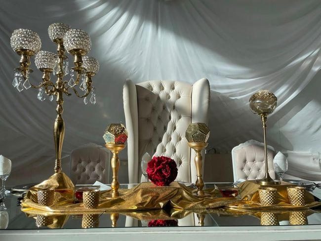 Elegante mesa dorada con candelabros de cristal, una rosa roja y sillas blancas capitoné sobre un fondo drapeado.