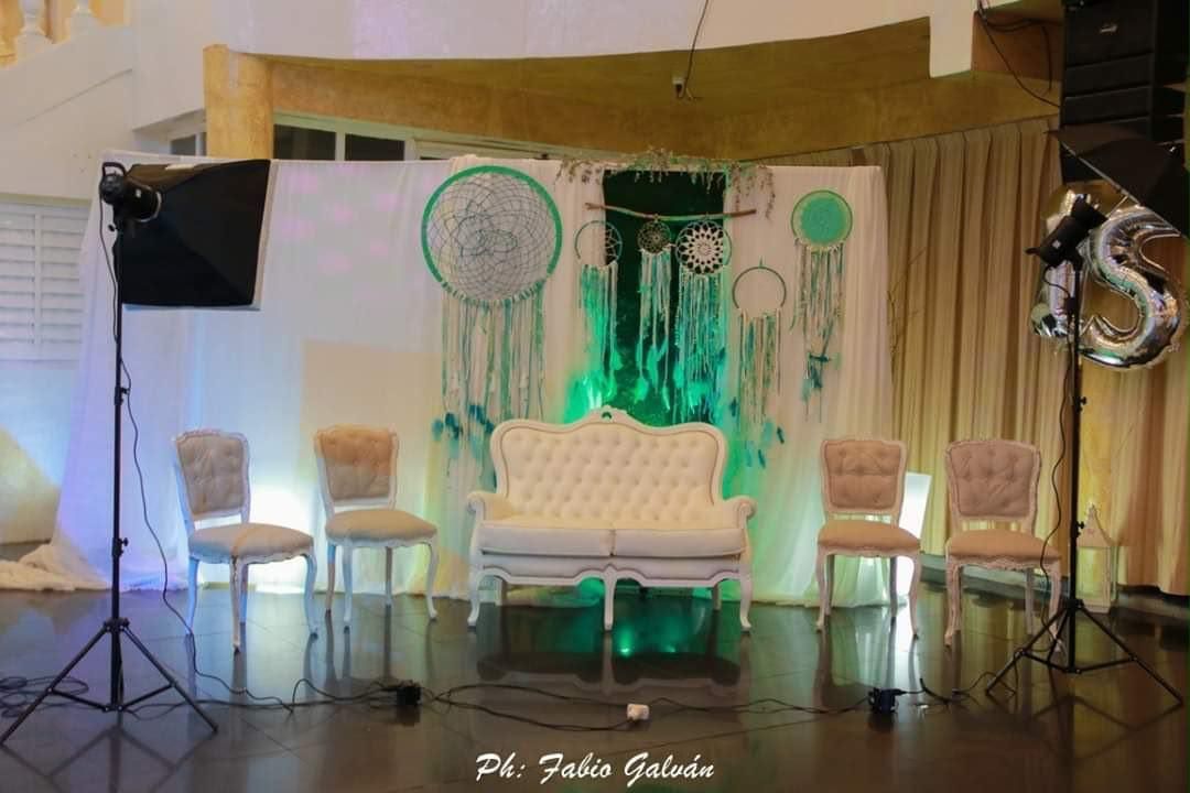 Escenario decorado con sofá de dos plazas blanco, sillas, iluminación verde y fondos para fotomatón.