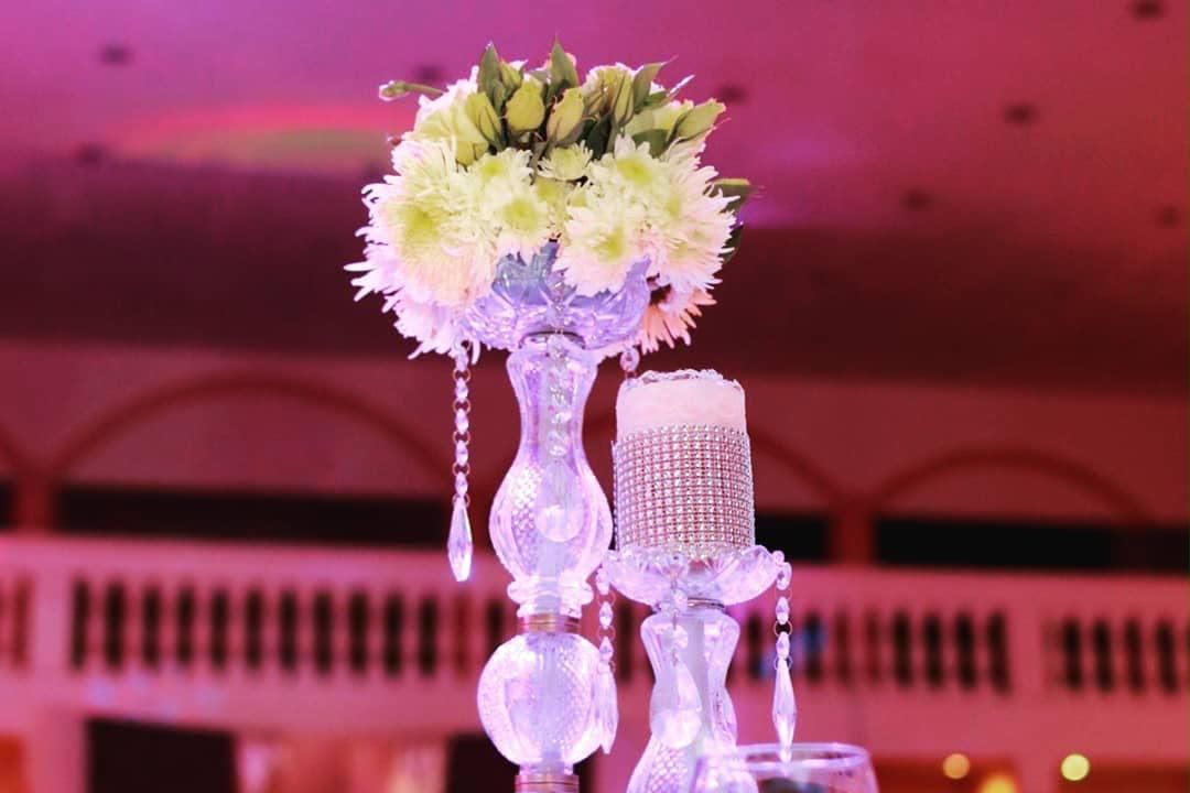 Centro de mesa floral sobre una mesa de banquete bajo iluminación rosa, con candelabros de cristal y cristales.