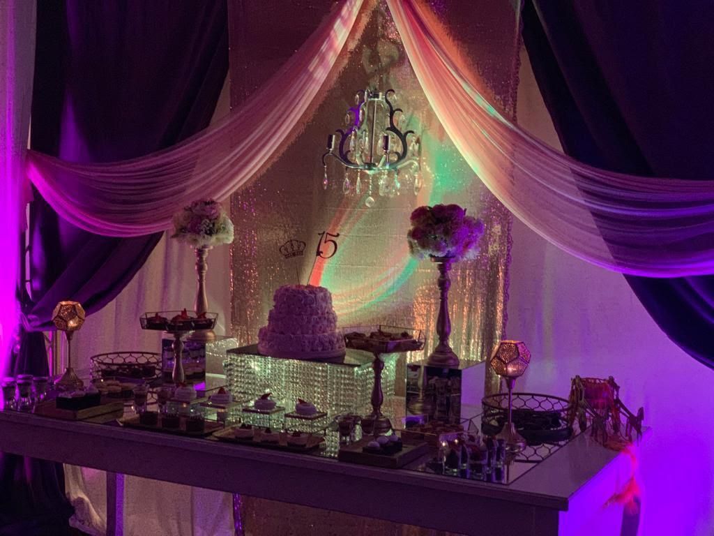 Mesa de postres para banquetes iluminada en color púrpura con tela drapeada, lámpara de araña y elegantes centros de mesa.
