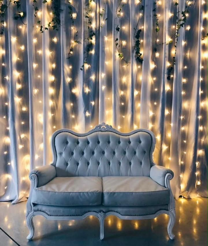 Sofá de dos plazas azul capitoné frente a cortinas blancas con luces de hadas cálidas
