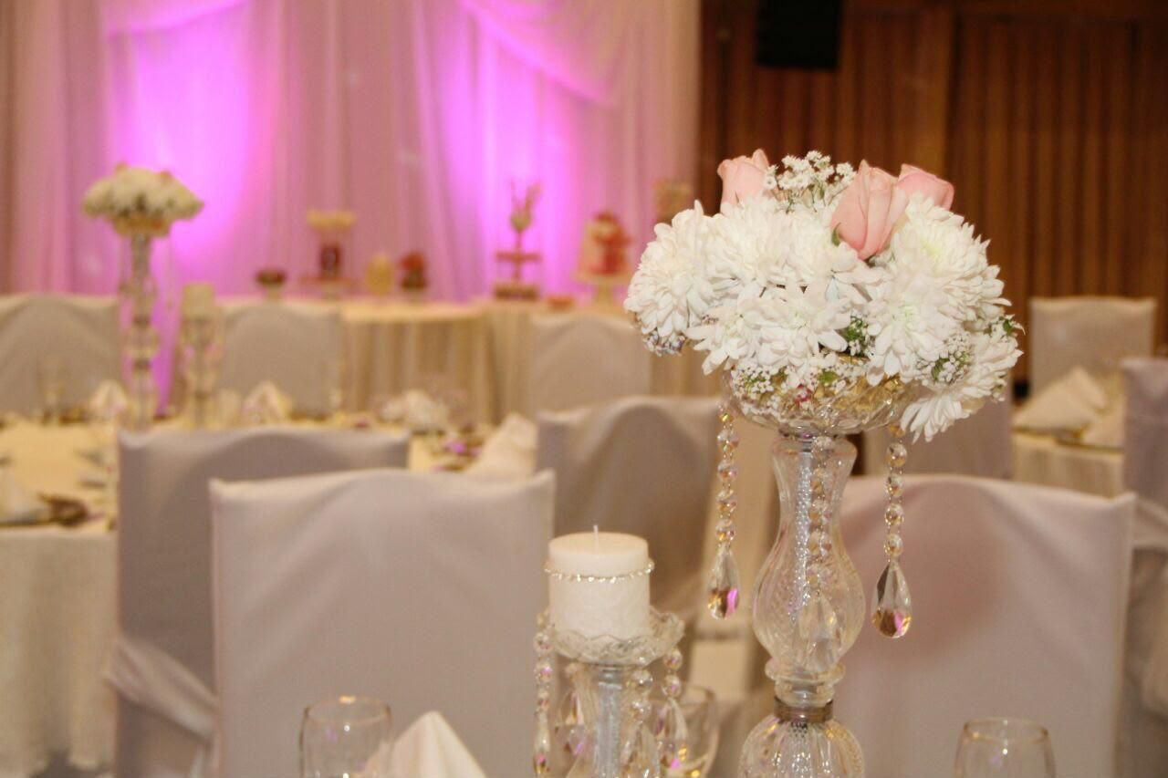 Elegante salón de banquetes con centros de mesa florales blancos e iluminación rosa tenue sobre mesas decoradas.