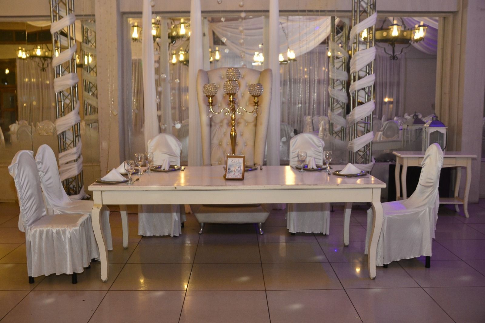 Elegante mesa de comedor decorada en una sala de eventos con iluminación tenue, sillas blancas y cortinas transparentes.