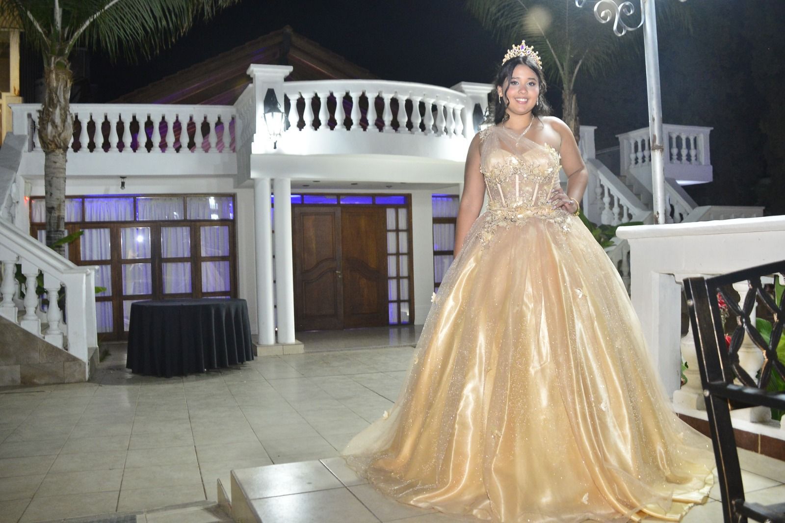 Mujer con vestido de gala dorado de pie en un patio iluminado por la noche.
