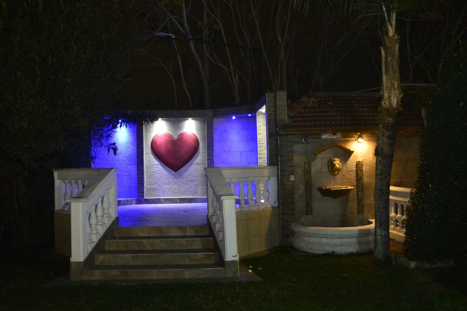 Entrada nocturna con una escultura de un corazón rojo, escalones iluminados y una pequeña fuente a la derecha.