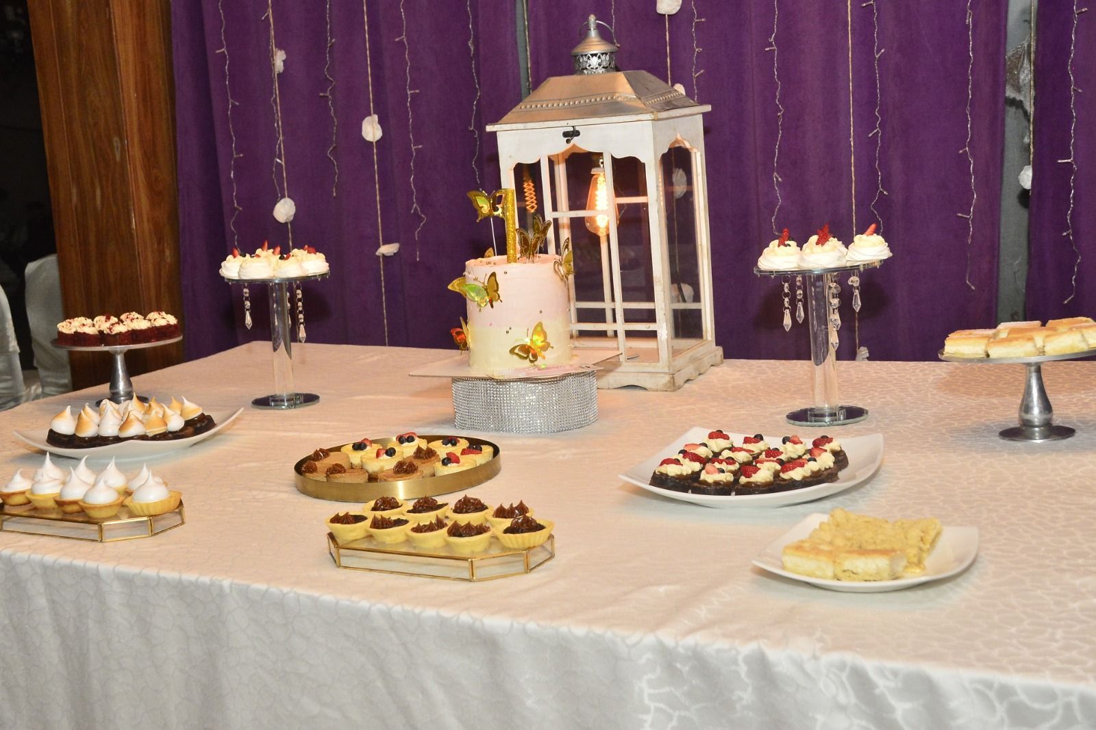 Mesa de postres con pasteles, tartas y un centro de mesa con una linterna blanca sobre un fondo morado.