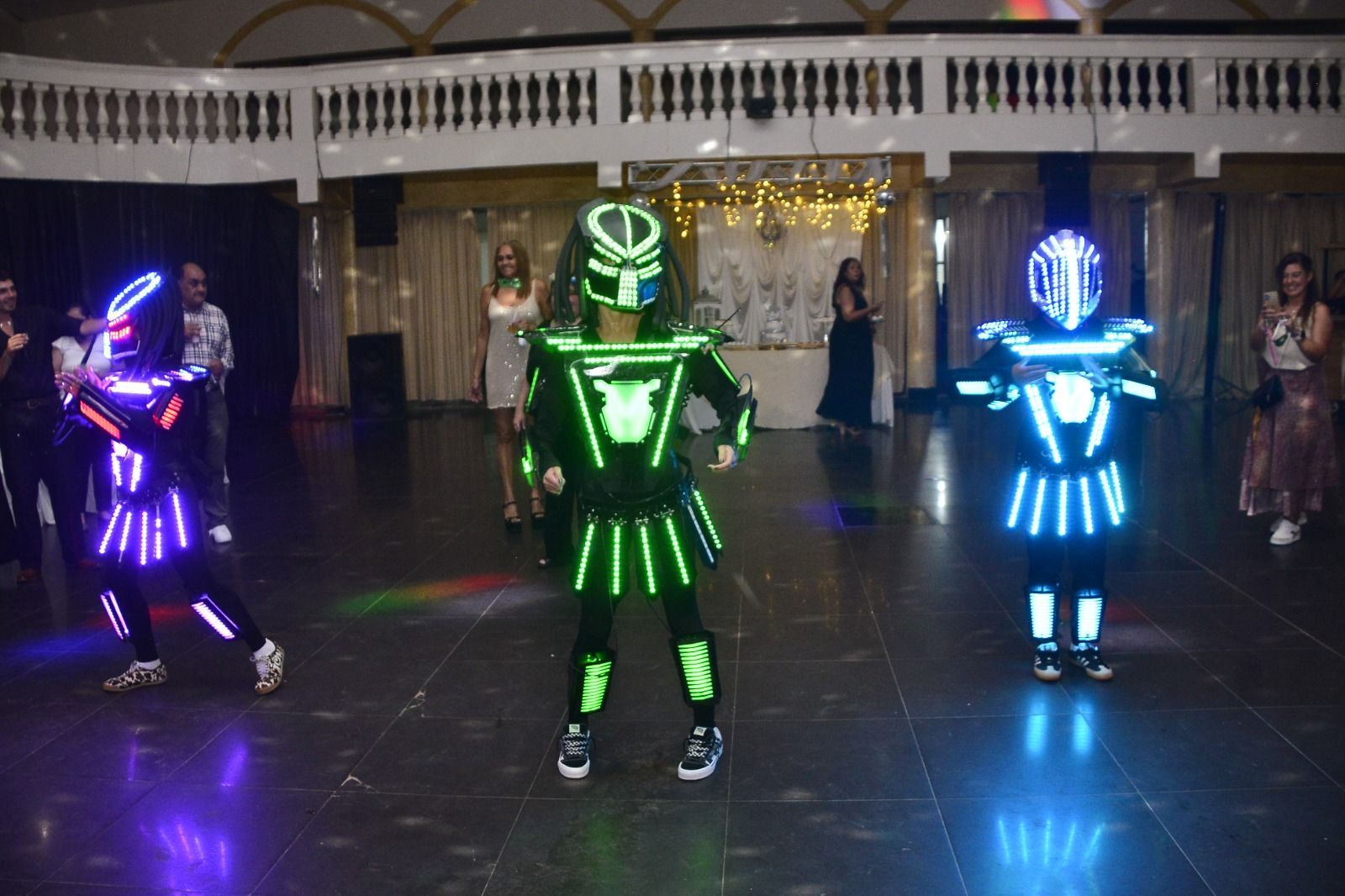 Tres artistas vestidos con trajes de neón, iluminados con luces LED, bailan sobre un suelo oscuro en un evento en interiores.
