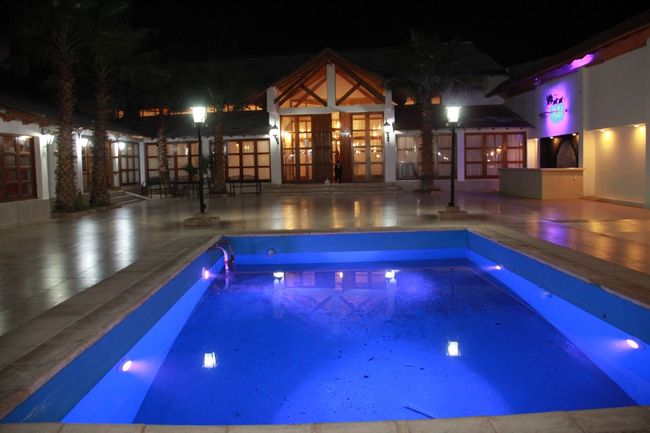Piscina cubierta iluminada con luz azul en un patio por la noche, con las luces del edificio reflejándose en la terraza.