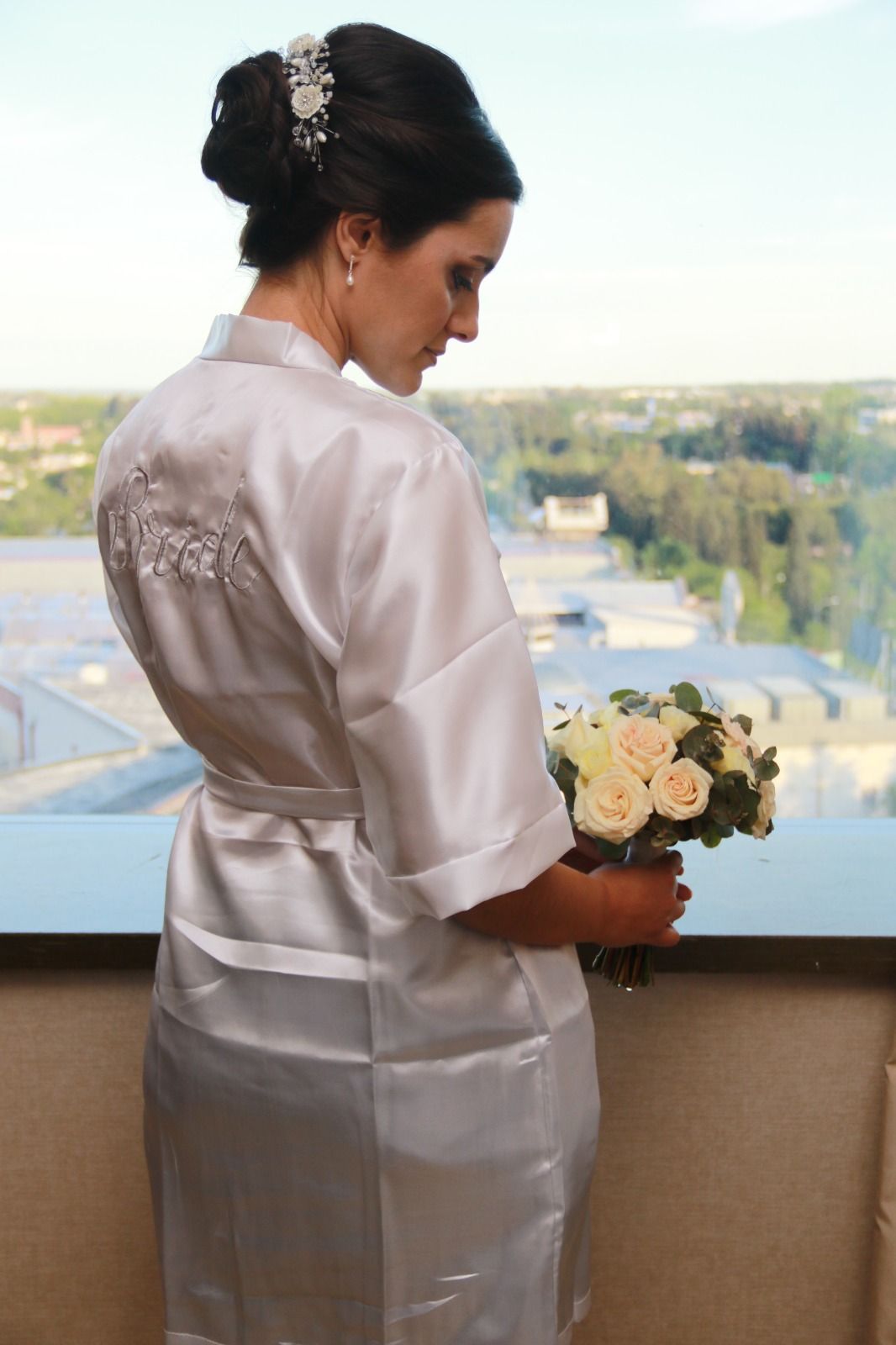 Mujer con túnica blanca sosteniendo un ramo de flores, de pie junto a una ventana con vista a la ciudad.
