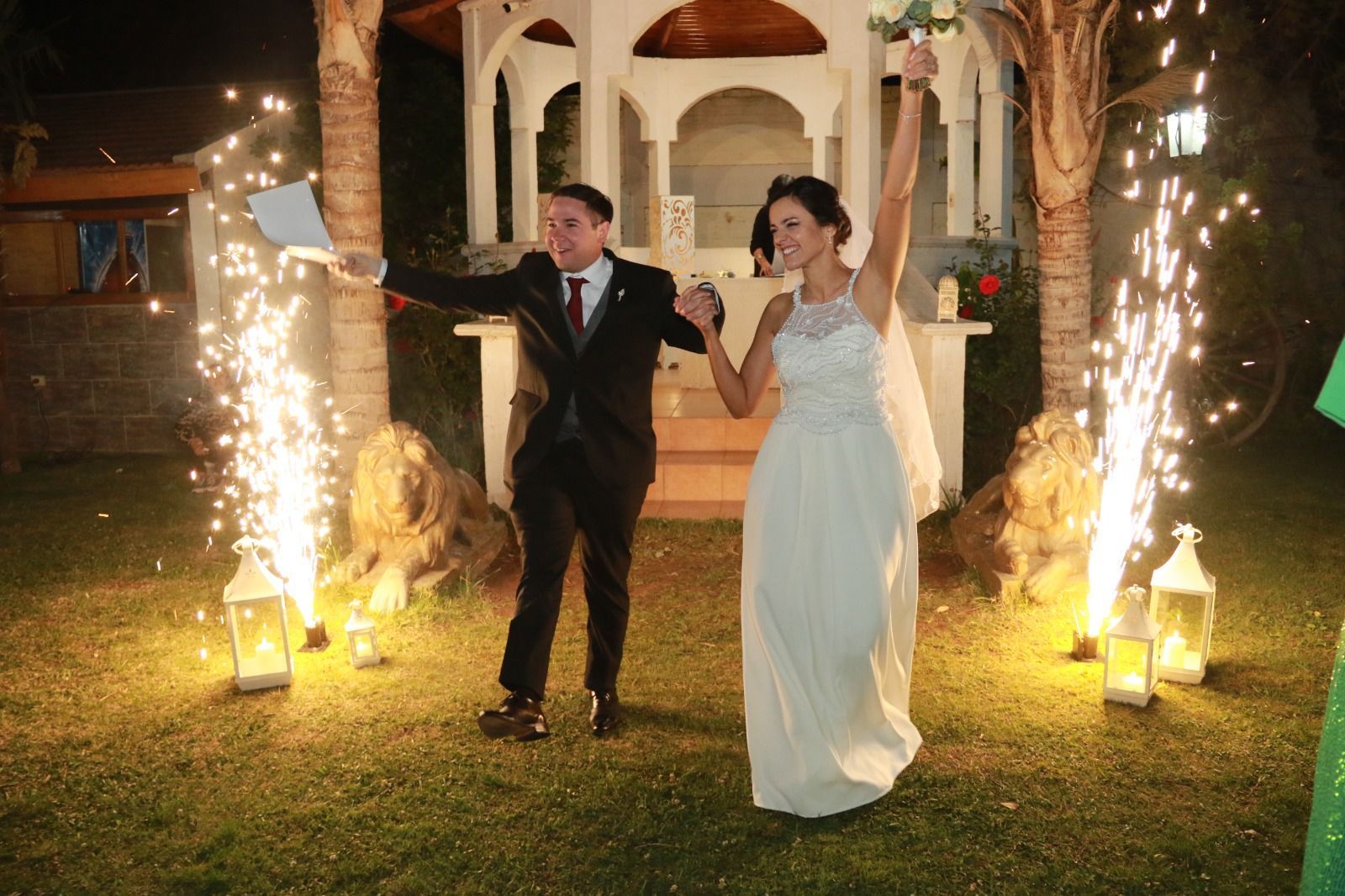 Una pareja vestida de gala camina entre fuegos artificiales en una celebración nocturna al aire libre.