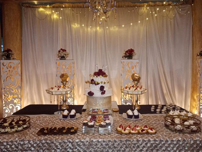 Escenario decorado con cortinas blancas, arreglos florales, velas y bandejas de dulces para una celebración.