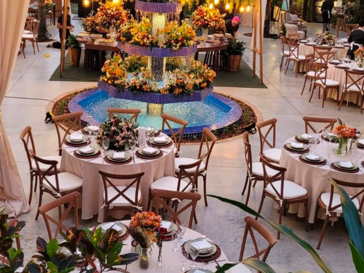 Elegante banquete ao ar livre com mesas redondas, arranjos florais e uma fonte iluminada em azul ao centro.