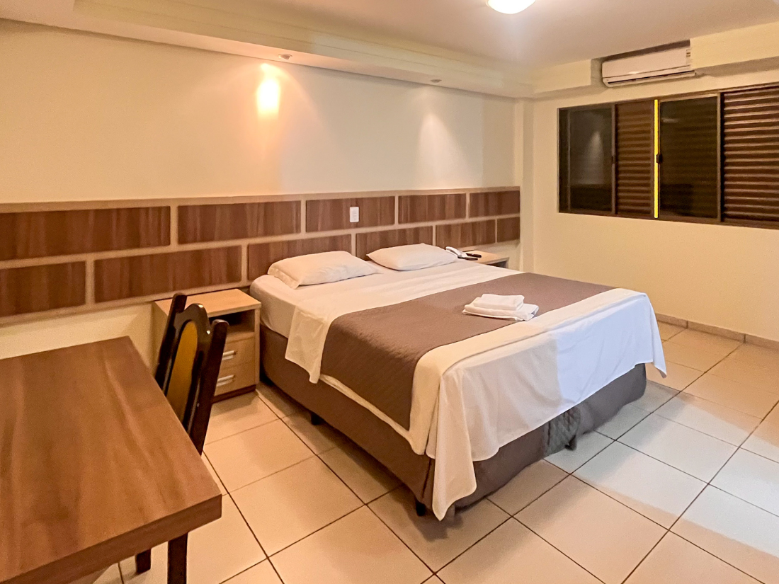 Quarto arrumado com cama de casal, escrivaninha de madeira, piso de azulejo e persianas.