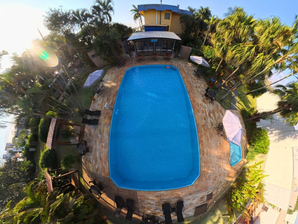 Vista aérea de um quintal com uma grande piscina azul rodeada por palmeiras e um terraço com mesas e cadeiras.