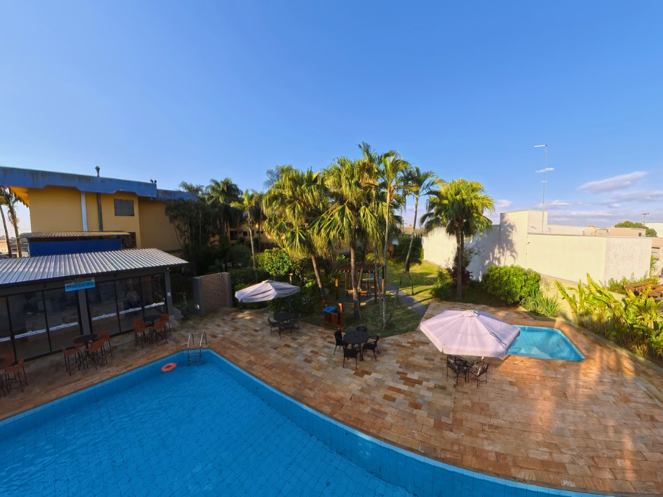 Quintal com piscina, palmeiras, guarda-sóis e uma casa sob um céu azul límpido.
