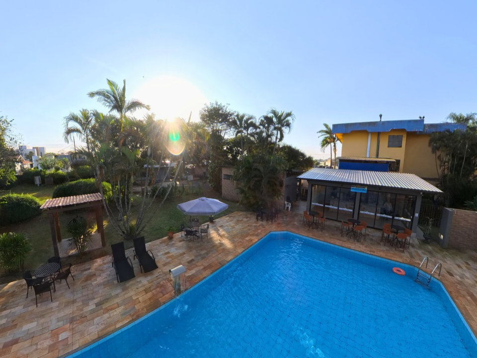 Piscina no quintal ao pôr do sol com palmeiras, pátio e uma casa de dois andares ao fundo.
