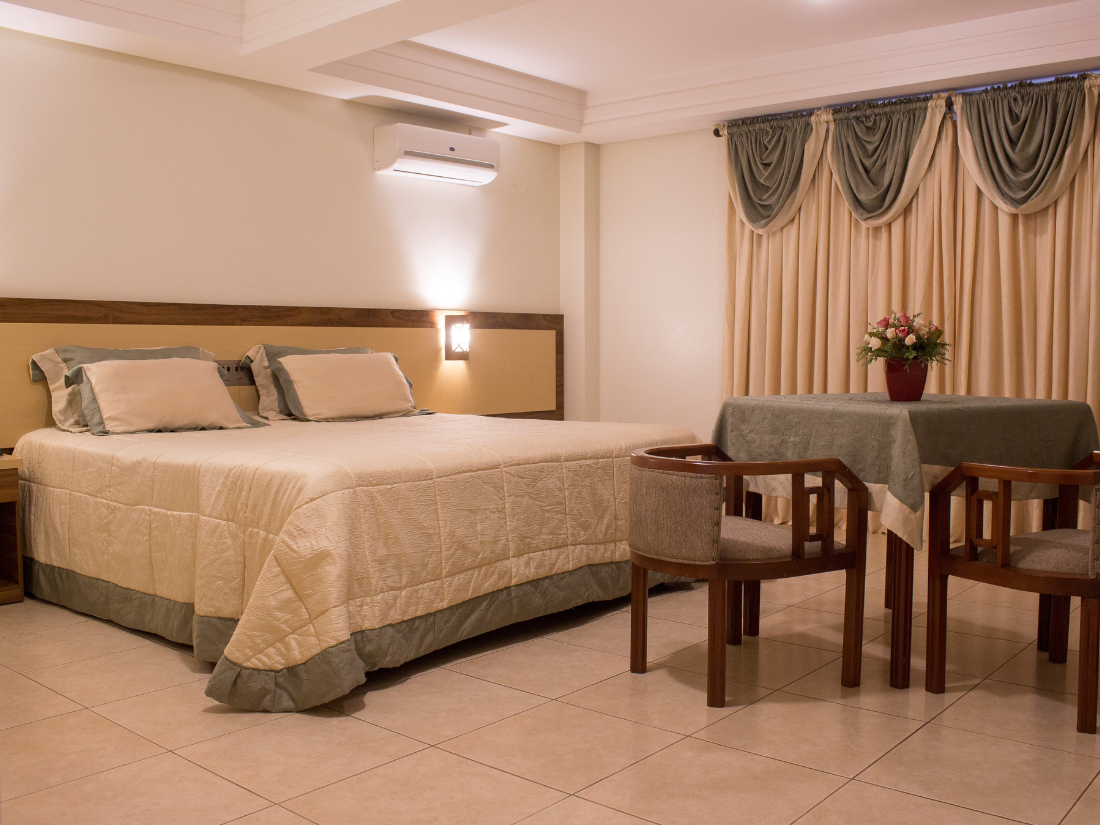 Quarto com cama de casal, mesa e cadeiras pequenas e cortinas drapeadas em um quarto bege aconchegante.
