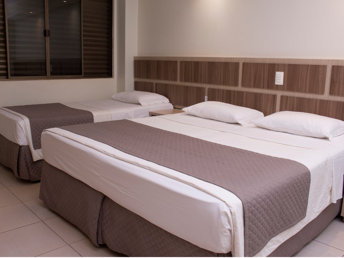 Quarto de hotel com duas camas bem arrumadas, lençóis brancos, colchas marrons e uma janela com persianas.