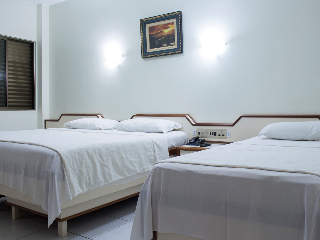 Quarto de hotel simples com três camas de solteiro bem arrumadas, paredes azul-claras e um quadro acima das cabeceiras.