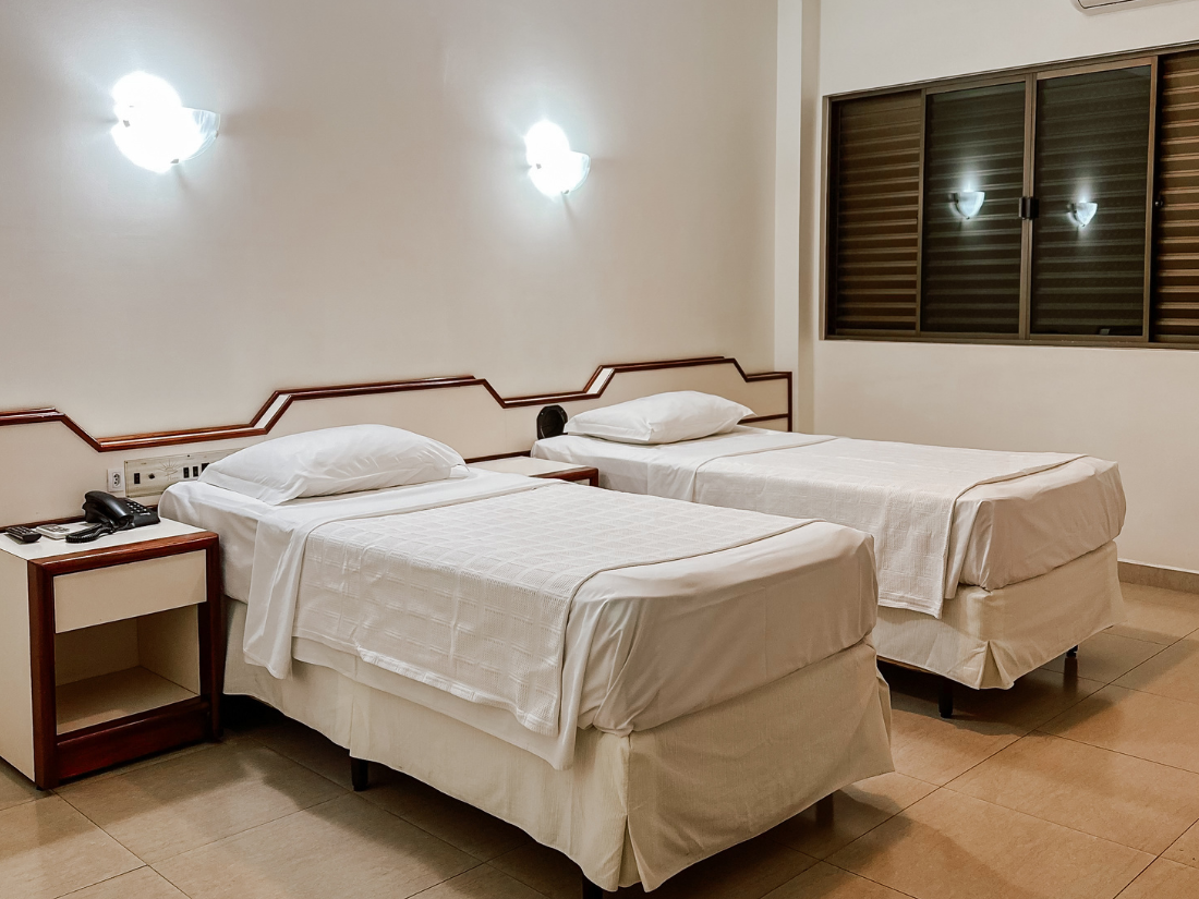 Um quarto de hotel arrumado com duas camas de solteiro, lençóis brancos, apliques de parede, uma mesa de cabeceira e uma janela.