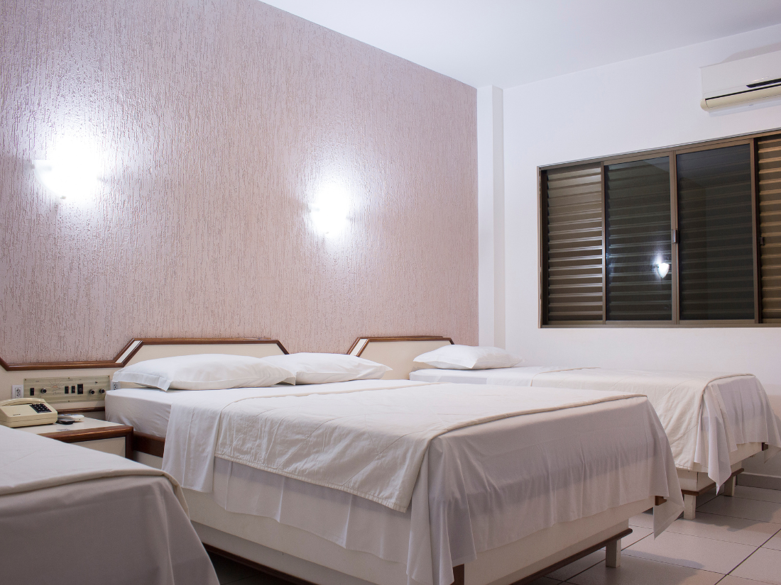 Quarto pequeno com três camas arrumadas, parede texturizada rosa e janela com persianas.
