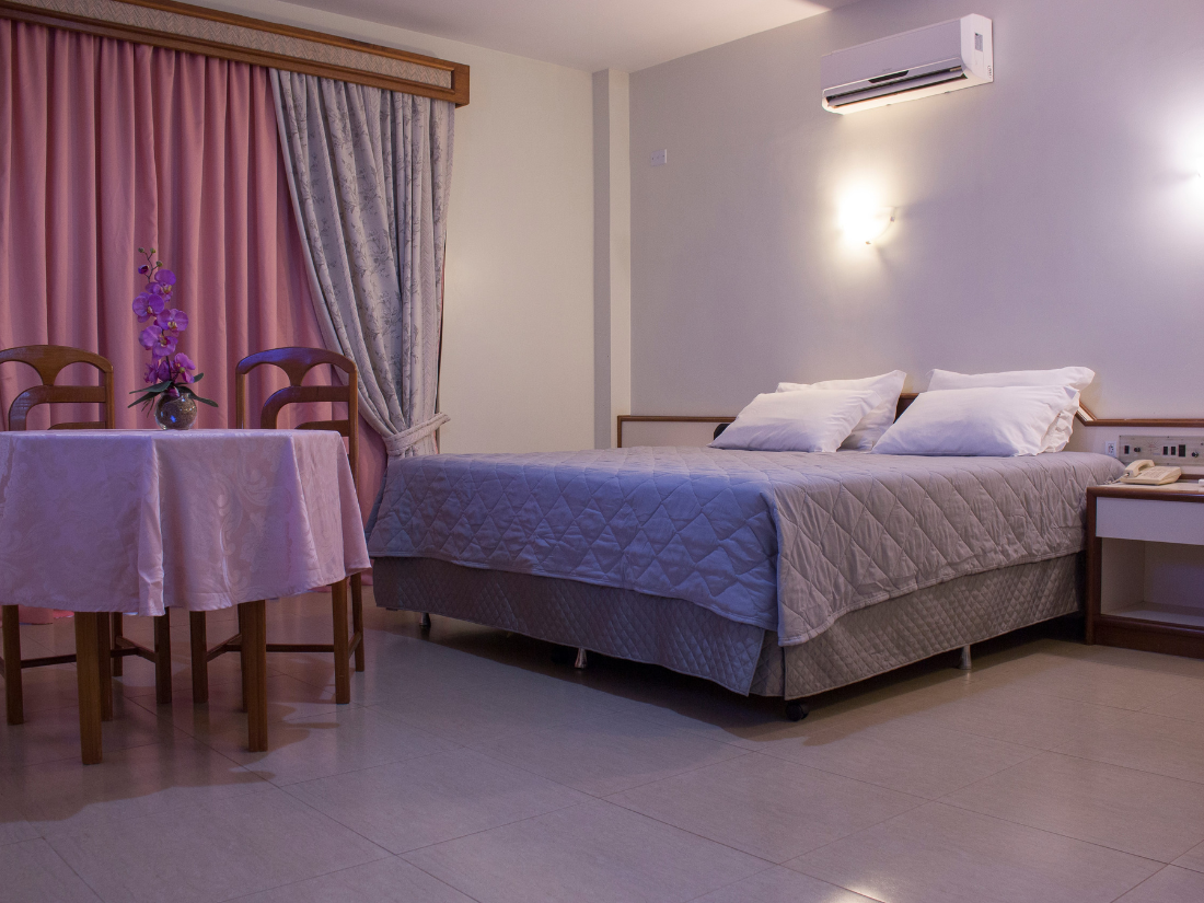 Quarto de hotel aconchegante com cama, mesa redonda, cadeiras, cortinas e apliques de parede em tons suaves de rosa e bege.